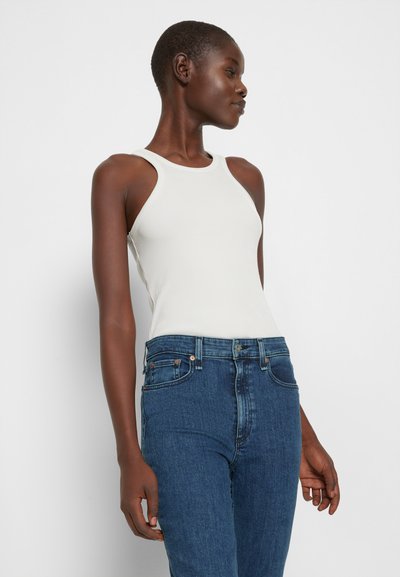 Top de alças largo branco com texturas, combinado com jeans de ganga escuros de cintura alta apresentando um design clássico de cinco bolsos.