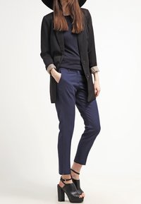 Svart kavaj med uppvikt ärm över en mörk tröja, marinblå skräddarsydda byxor och svarta blockklackssandaler. Enkelt, elegant design.