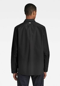 Camisa negra de manga larga con cuello abotonado, textura suave, con un sutil logotipo en la parte posterior y puños abotonados.