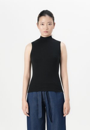 Femme avec les cheveux attachés, portant un haut sans manches noir à col montant et un pantalon en denim bleu à jambes larges avec une ceinture en tissu.