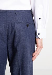 Marineblaue, maßgeschneiderte Hosen mit strukturierter Oberfläche, ausgestattet mit einer Gesäßtasche, die mit einem runden schwarzen Knopf gesichert ist, und einem sichtbaren Bund.