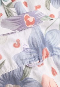 Blusa floreale realizzata in un tessuto leggero con fiori blu e corallo, foglie verdi e un accento lavanda sul colletto. Dettaglio di cucitura viola.