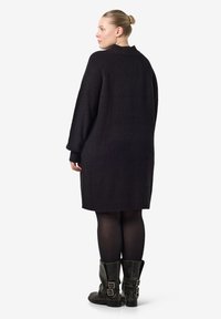 Μαύρο υπερμεγέθες πλεκτό φόρεμα με ραβδωτή υφή, μακριά μανίκια και στρογγυλή λαιμόκοψη, συνδυασμένο με μαύρες μποτές μέχρι τον αστράγαλο με λεπτομέρειες από πόρπες.