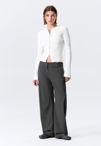Cardigan blanc côtelé avec une fermeture éclair à l'avant, associé à un pantalon large gris foncé. La tenue présente une coupe courte et des manches longues.