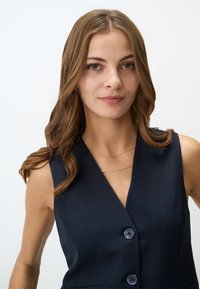 Navy ärmelloser Blazer mit tiefem V-Ausschnitt, ausgestattet mit zwei Knöpfen und einer taillierten Passform. Das Model trägt gestapelte goldene Halsketten, die Haare sind in sanften Wellen gestylt.