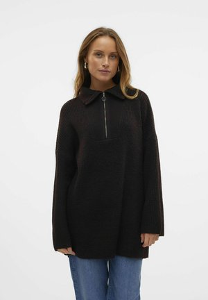 Pull noir en mélange de laine avec un col haut zippé, coupe ample, texture côtelée et manches longues, associé à un jean bleu.
