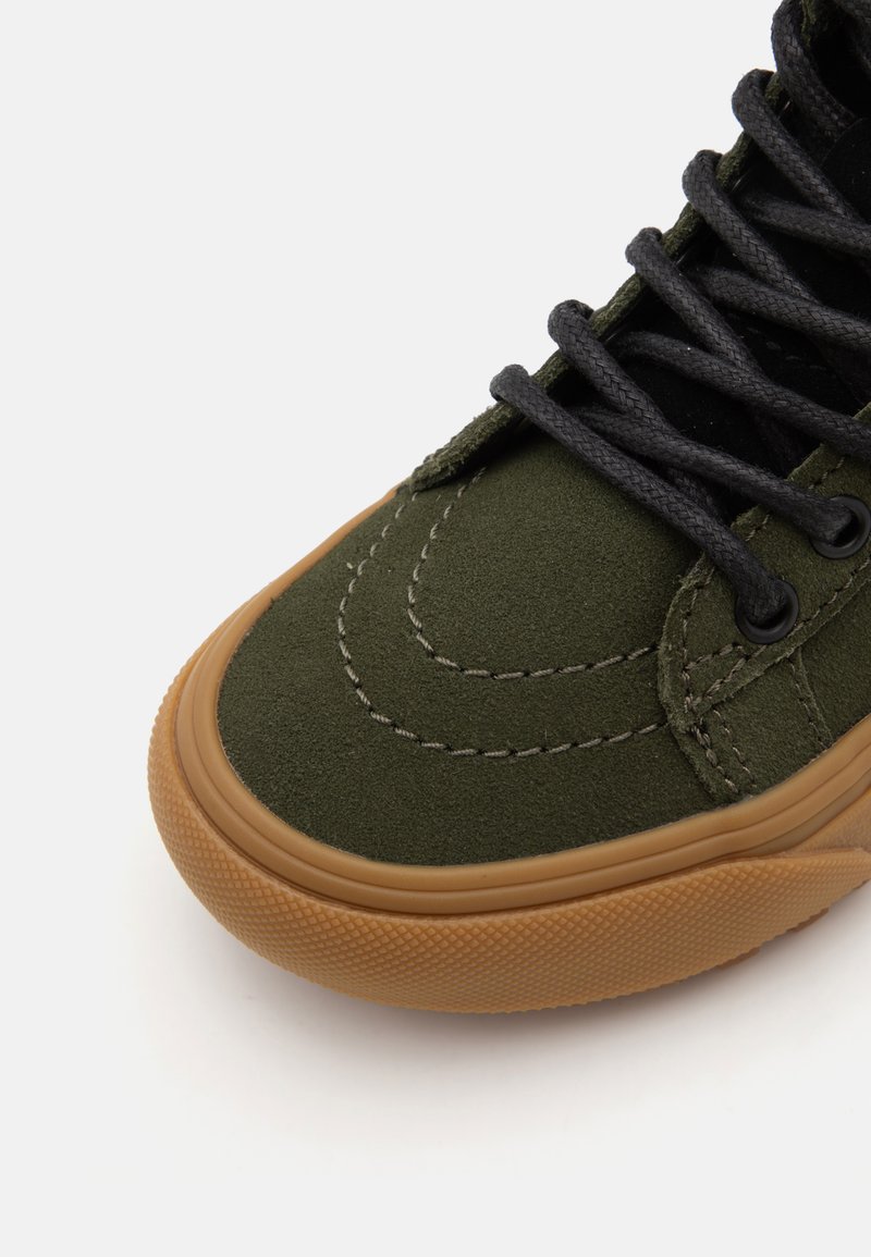 Zapatilla de ante verde con cordones negros, que presenta un detallado acolchado y una suela de goma marrón. La puntera redondeada y los acentos texturizados mejoran la agarre.