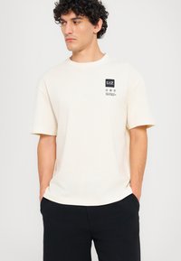 Cream-colored short-sleeve t-shirt with a round neckline and a black graphic label on the chest, paired with black pants. Soft texture.
