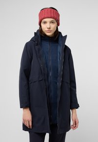Marineblauwe waterdichte jas met een ritssluiting en drukknoopsluitingen, gelaagd over een blauwe fleece top, aangevuld met een roze gebreide haarband.
