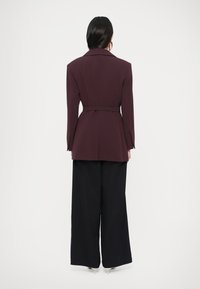 Bordó testre szabott blézer övvel a derekán, bevágott hajtókával és strukturált vállakkal, széles szárú fekete nadrággal párosítva.