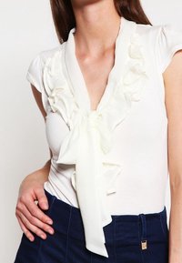 Bílá blůza s volánkovým výstřihem a detailem mašle. Krátké rukávy typu cap a měkká, hladká látka. Vpředu v kombinaci s tmavě modrými kalhotami s vysokým pasem.