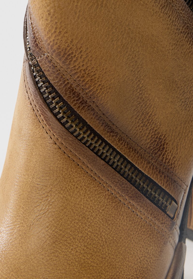 Gros plan sur une botte en cuir marron montrant la texture, les détails de couture et une fermeture éclair en métal noir diagonale.
