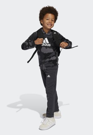 Felpa nera a fantasia mimetica con logo "Adidas" bianco, pantaloni grigi e sneakers chiare. Un bambino posa con uno zaino, sorridendo.