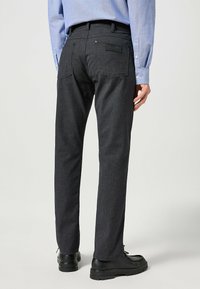 Donkergrijze slim-fit jeans met een gladde textuur, vijfzakdesign en een merkpatch op de achterkant, gecombineerd met zwarte schoenen.