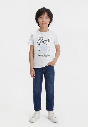 Otrok s črnimi lasmi, oblečen v belo majico Guess, modre jeans hlače in bele superge, stoji z roko v žepu.