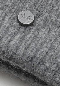 Tessuto grigio a maglia con finitura testurizzata, caratterizzato da un bottone metallico circolare inciso con la scritta "PEARLWOOD" e linee decorative.