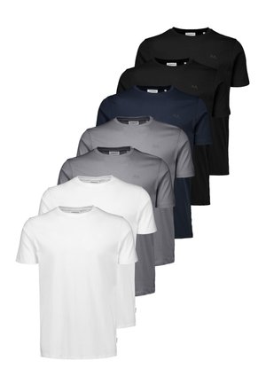Jonge man met lichtbruin haar staat met het gezicht naar voren, draagt een effen zwart T-shirt en blauwe jeans tegen een neutrale achtergrond.