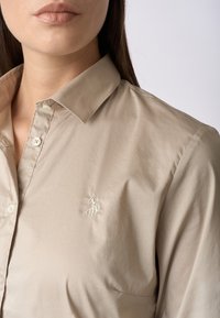 Camicia beige con bottoni, realizzata in tessuto morbido, dotata di colletto a punta e logo ricamato sul lato sinistro del petto. Finitura texturizzata ma elegante.