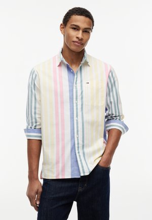 Ung mand iført pastel flerfarvet skjorte med lodrette striber og optrukne ærmer samt mørke jeans, poserer med den ene hånd i lommen.