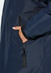 Chaqueta impermeable de color azul marino con un bolsillo lateral con cremallera, puños elásticos y una tela lisa y texturizada. El tirador de la cremallera tiene una etiqueta de tela.