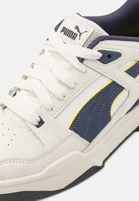 Sneaker bianca in suede e pelle con accenti blu navy, dettagli gialli di lato e lacci testurizzati. Logo PUMA sulla linguetta.