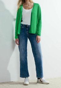 Cardigan leggero verde sopra a una maglietta bianca, abbinato a jeans a gamba larga blu chiaro e sneaker bianche con accenti rossi.