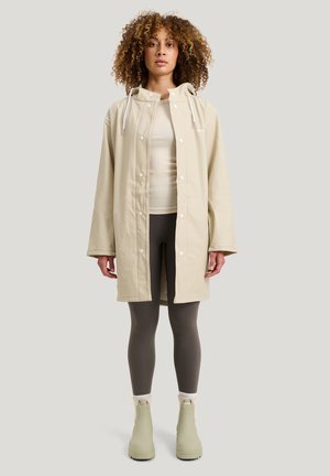 Beige vanntett jakke med trykknapper og snor i hetten, kombinert med en lys topp og grå tights, iført grønne ankelstøvler.