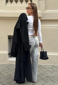 Svart lång jacka hängande över axeln, vit skräddarsydd långärmad skjorta, ljusgrå jeans och en texturerad svart vävd handväska. Sido vy, stadsmiljö.