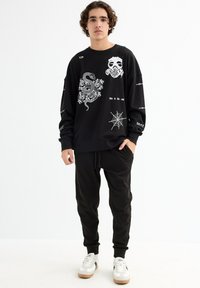 Sudadera negra con estampados gráficos blancos, que presenta una serpiente, una máscara de calavera y texto; combinada con pantalones jogger negros y zapatillas blancas.