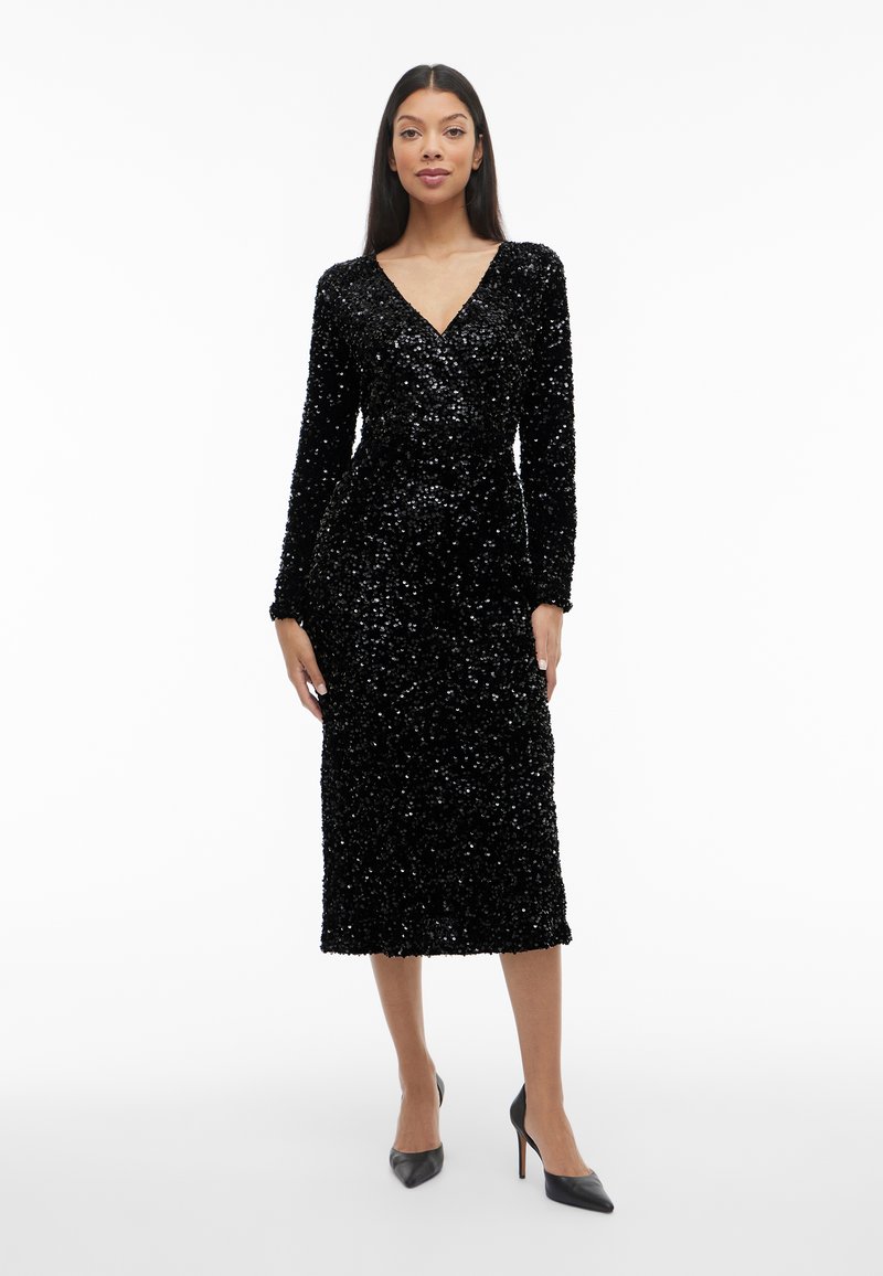 Sieviete, tērpusies garām piedurknēm, melnā spīdīgu secināto midi kleitā ar V veida kakla izgriezumu un melnām augstpapēžu kurpēm, stāv uz balta fona.