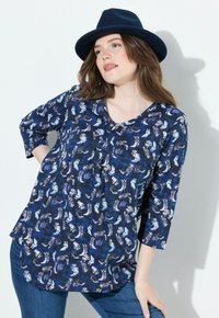 Mujer con un sombrero azul marino y una blusa de tres cuartos con estampado floral azul, posando frente a un fondo blanco.
