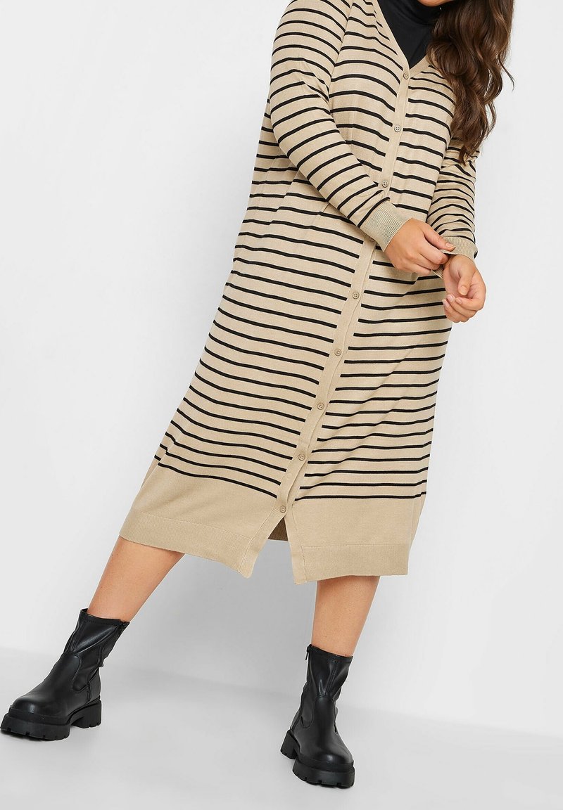 Béžový pletený kardigan s černými pruhy, výstřihem do V, knoflíky vpředu a bočními rozparky. Nošený s černými kotníkovými botami, vytváří vrstvený vzhled.