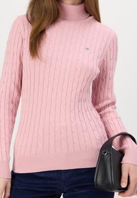 Pull à col roulé en maille côtelée rose avec un motif torsadé, silhouette ajustée et ourlet côtelé. Associé à un petit sac à main noir.