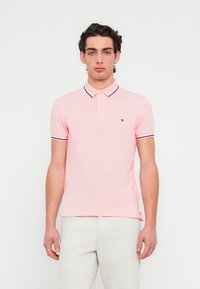 Πόλο μπλουζάκι με κοντά μανίκια σε ανοιχτό ροζ βαμβάκι, με γιακά δύο κουμπιών, αντίθετες ναυτικές ρίγες στα μανίκια και μικρό λογότυπο στο στήθος.