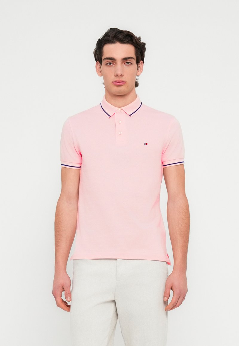 Πόλο μπλουζάκι με κοντά μανίκια σε ανοιχτό ροζ βαμβάκι, με γιακά δύο κουμπιών, αντίθετες ναυτικές ρίγες στα μανίκια και μικρό λογότυπο στο στήθος.