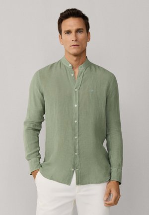 Bărbat care poartă o cămașă verde deschis cu mâneci lungi, nasturi și guler mandarin, asortată cu pantaloni albi, stând într-un decor neutru.