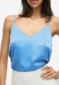 Svetlo modrý saténový top na ramienkach s výstrihom do V, tenkými ramienkami a voľným strihom, skombinovaný s bielou sukňou s textúrovaním a nariasením.