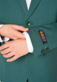 Blazer verde su misura con due bottoni, caratterizzato da una toppa ricamata "DAILY" e un'accenno arancione. Polsino della camicia bianca visibile sotto.