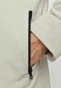 Giacca beige chiaro con una tasca con zip nera. Texture liscia, design aderente. Cuciture dettagliate lungo il bordo, che mostrano uno stile moderno.