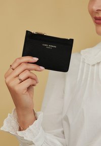 Black textured leather cardholder with a gold zipper and embossed brand name. Hand holding cardholder against a plain background.