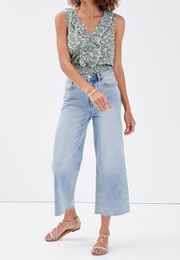 Haut sans manches vert à motif floral avec un col en V et un bord à volants, porté avec un jean large taille haute délavé clair et des sandales beige.