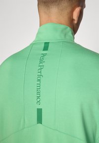 Der grüne Viertelreißverschluss-Pullover verfügt über einen hohen Kragen, Flachnähte und ein auffälliges „Peak Performance“-Branding vertikal auf der Rückseite. Weicher Stoff.