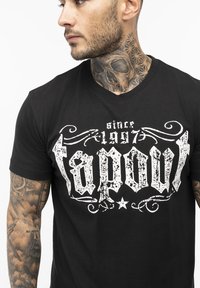 Fekete pamut póló, melyen egy nagy, megkopott fehér "tapout" grafika található, díszítő motívumokkal és a fölötte lévő "1997 óta" felirattal.