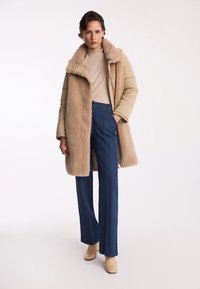 Beżowa kurtka puchowa z futrzanym kołnierzem, jasnobeżowy sweter, granatowe spodnie o szerokich nogawkach oraz beżowe botki na obcasie.