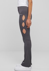 Persona valkājot ribotas tumši pelēkas bikses ar trim dimanta formas izgriezumiem uz augšstilba, baltas sneakers un saspringtu baltu blūzi.