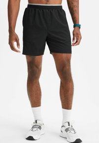 Schwarze Sportshorts mit einem elastischen Bund und einer glatten Textur, oberhalb des Knies geschnitten, kombiniert mit weißen Socken und Sportschuhen.