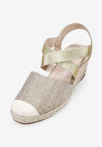 Wedge sandal with a beige fabric upper, gold metallic cross-straps, woven jute sole, and a white braided toe cap.