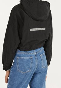 Black hooded jacket in lightweight fabric, featuring a reflective strip across the back; paired with light blue denim jeans.