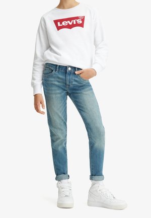 Personne portant un sweat-shirt blanc Levi's, un jean bleu clair retroussé et des baskets montantes blanches, debout avec une main dans la poche.