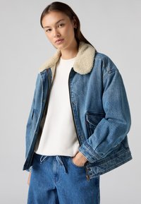 Veste en denim bleu clair avec un col beige et duveteux. Elle possède de grandes poches et une fermeture éclair à l'avant. Associée avec un haut blanc et un jean bleu.
